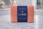 Packaged coral pink Vorre Norvelle recycled cotton bath towel neatly folded and wrapped with a signature dark blue brand paper band.